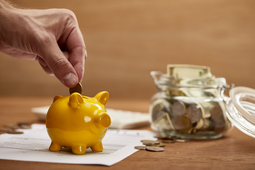 man placing a penny in small yellow piggy bank, jar of coins sitting nearby